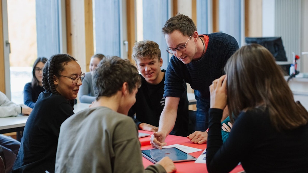 Fünf Schülerinnen und Schüler sind mit einem Tablet, auf welchem ihnen der Lehrer etwas erklärt, um einen Tisch versammelt.