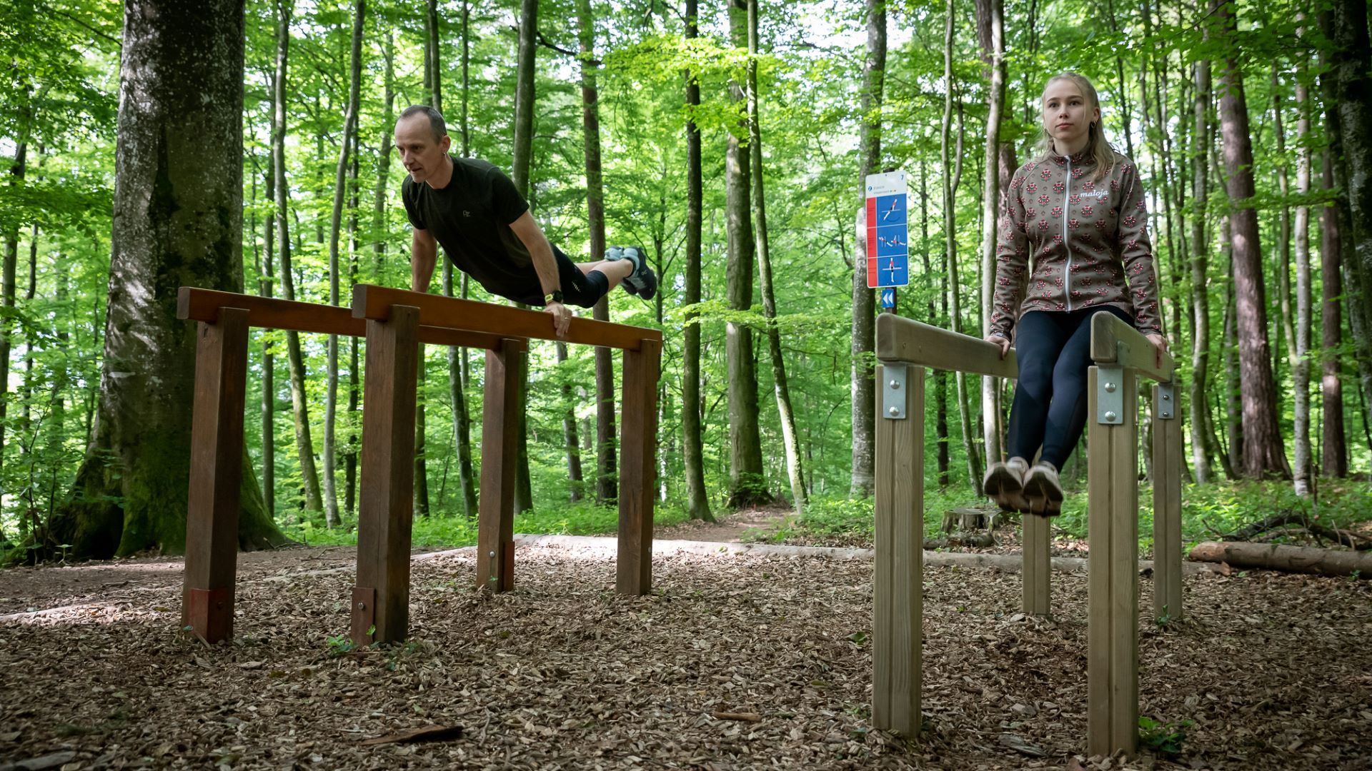 Two individuals are doing gymnastics in the forest on parallel bars, and in the background, you can see the board with exercise instructions.