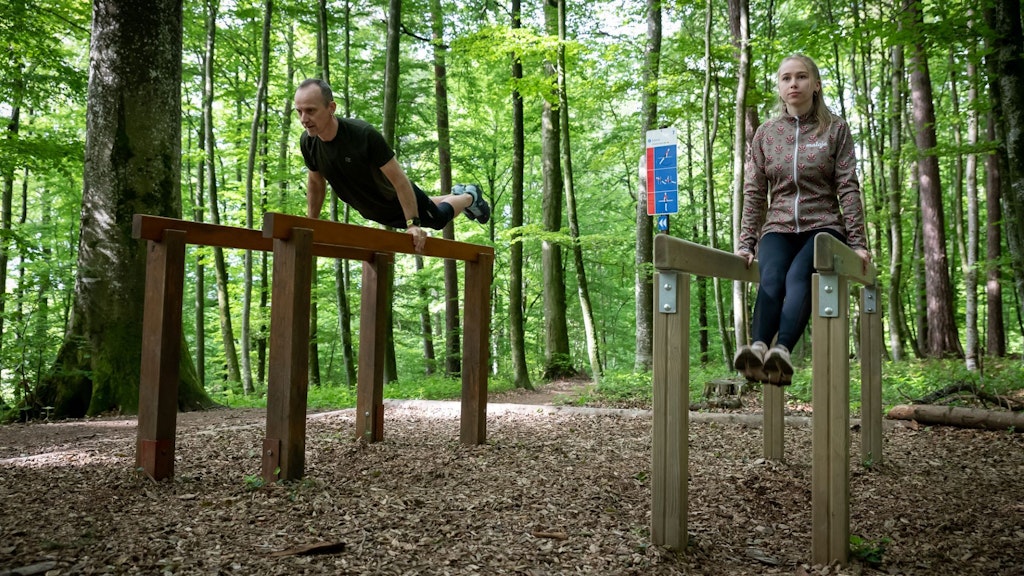 Deux personnes font de la gymnastique dans la forêt sur des barres parallèles, et à l’arrière-plan, on voit le panneau avec les instructions d’exercice.