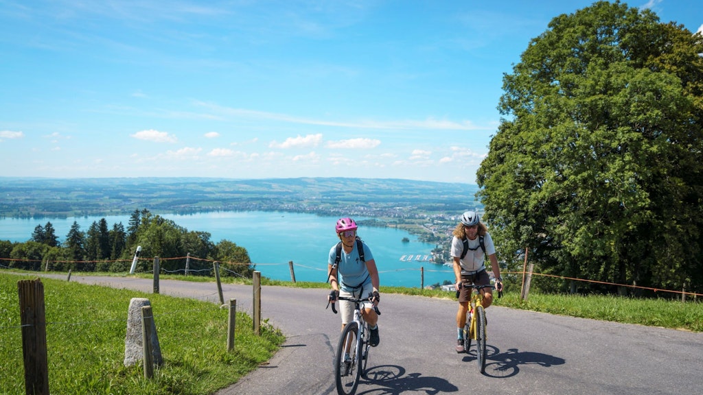 Deux cyclistes sur une route près d’un arbre, avec un lac et des collines en arrière-plan.