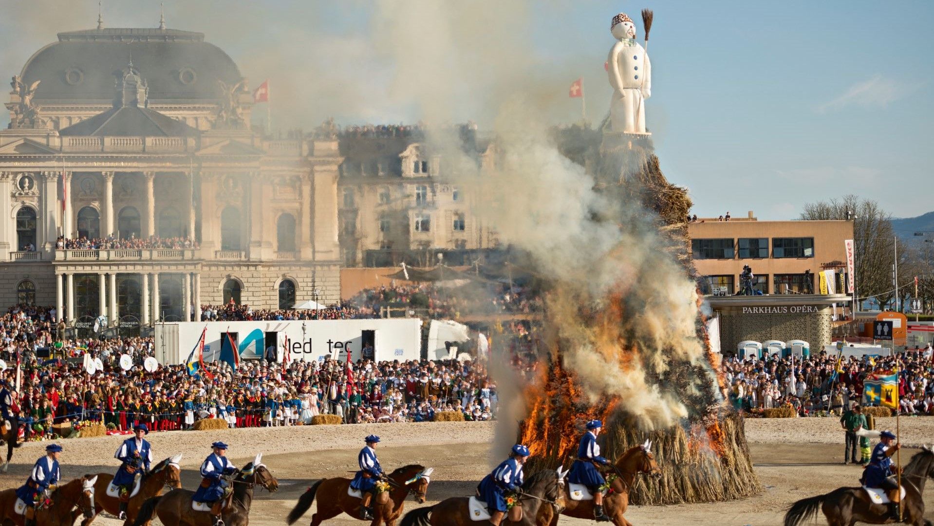 Cavaliers en tenue traditionnelle galopant devant un bûcher en feu avec un bonhomme blanc au sommet, dégageant de la fumée, lors d'un événement en plein air.