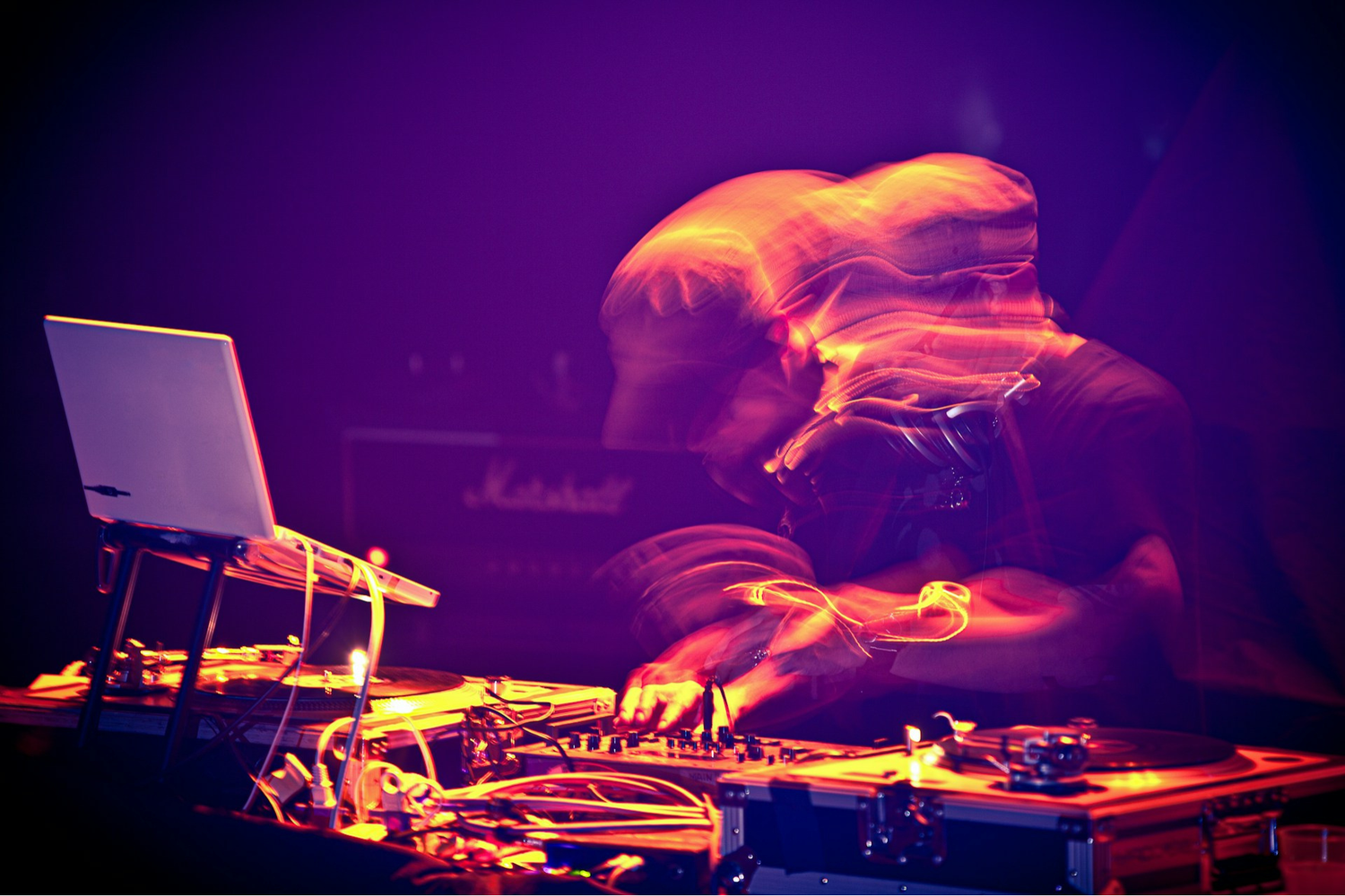 DJ performing live with laptop, turntables and mixer, lit by colourful lights with motion blur effect.