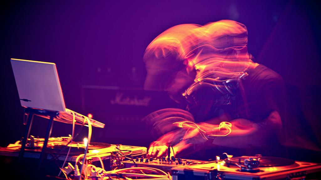 DJ performing live with laptop, turntables and mixer, lit by colourful lights with motion blur effect.