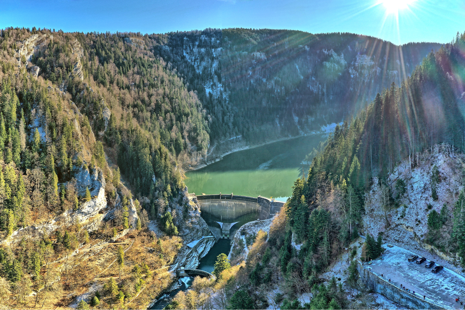 Aerial view of a curved dam in a narrow, wooded gorge, with a green reservoir behind it, steep rocky cliffs and a bright sun glinting on the right.