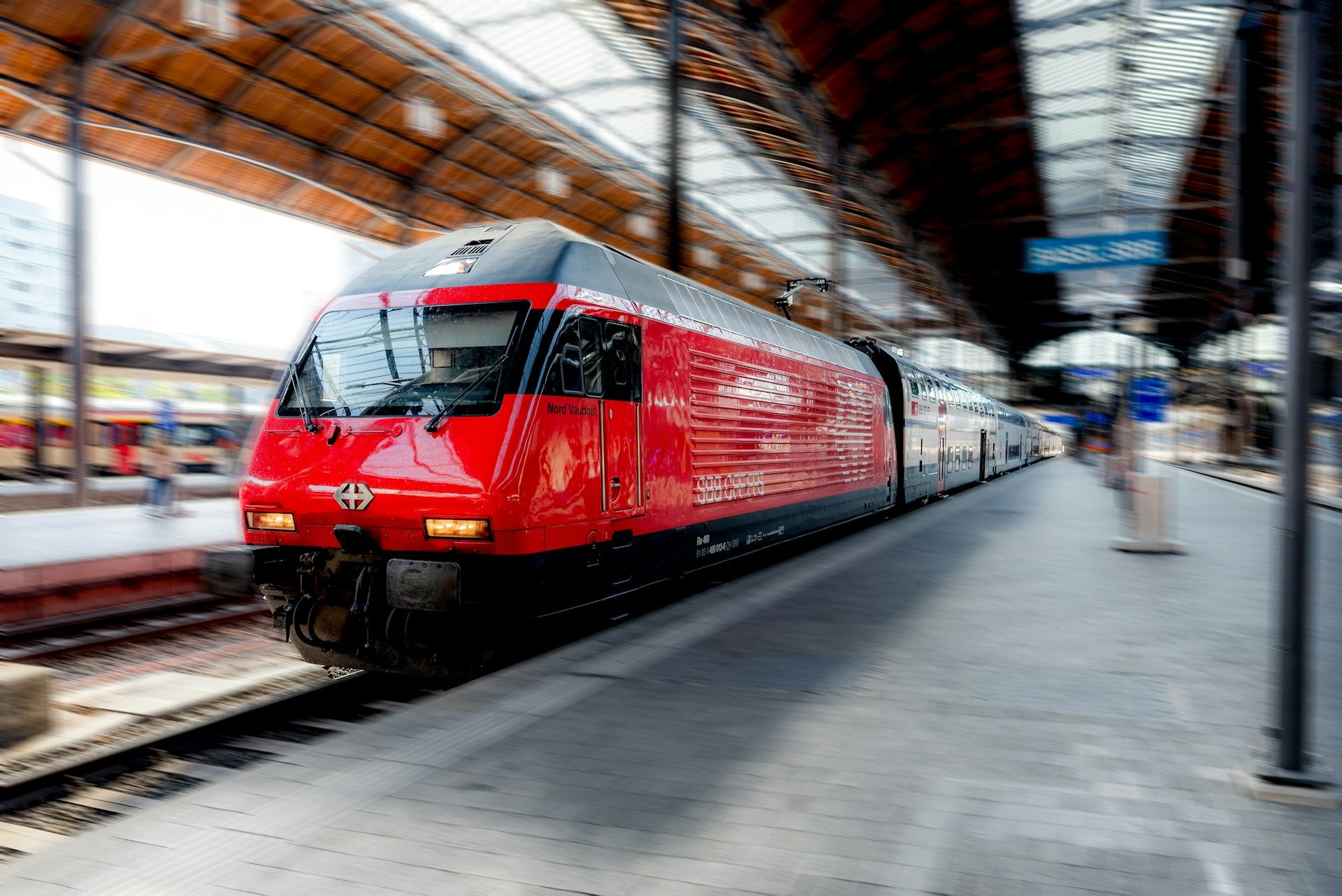 Train rouge CFF sur un quai de gare couvert, locomotive à l’avant en léger flou de mouvement, voûte en bois et verre au-dessus du quai.