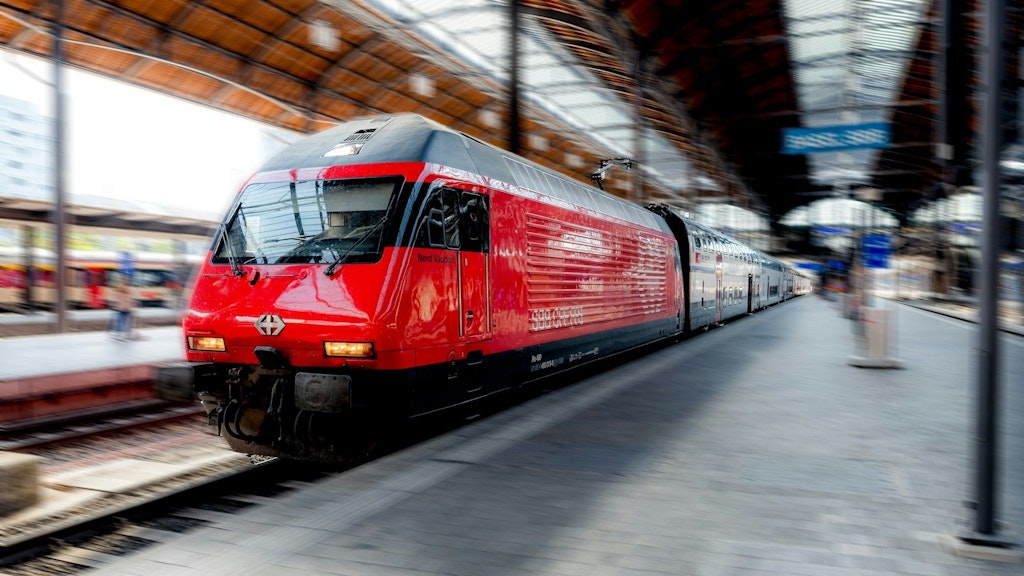 Train rouge CFF sur un quai de gare couvert, locomotive à l’avant en léger flou de mouvement, voûte en bois et verre au-dessus du quai.