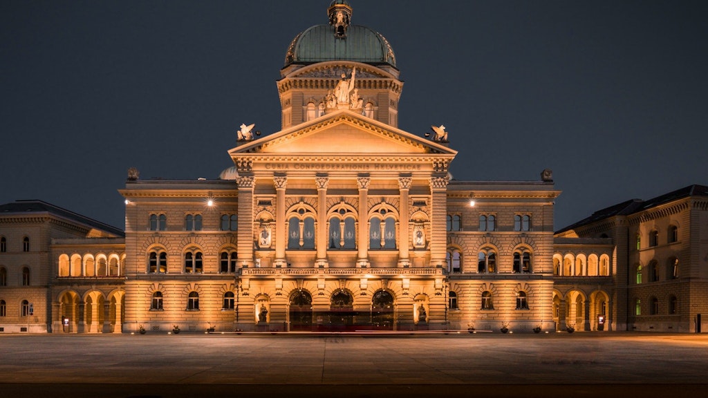 Vue de nuit du Palais fédéral suisse à Berne, éclairé, présentant sa façade symétrique en pierre avec arcades, colonnes et coupole, devant une vaste esplanade vide. 
