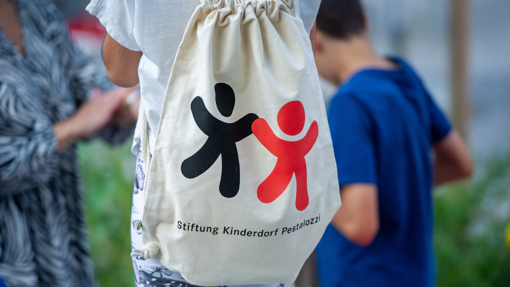 Canvas drawstring bag worn by a child, printed with a logo of two stylised black and red figures holding hands and the text ‘Stiftung Kinderdorf Pestalozzi’. 