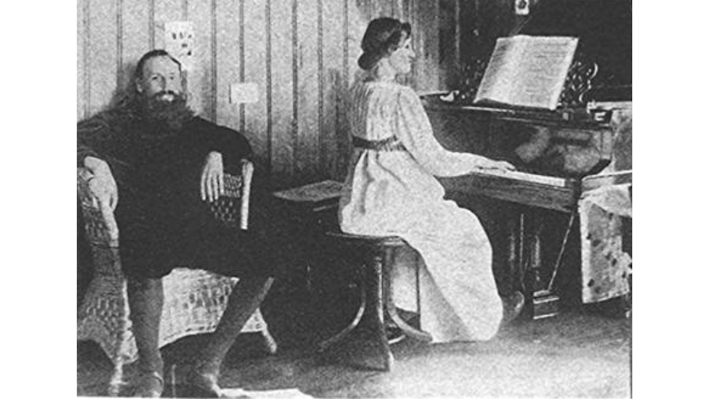 Fotografia storica con un uomo barbuto seduto su una sedia di vimini accanto a una donna che suona il pianoforte in una stanza rivestita in legno. 