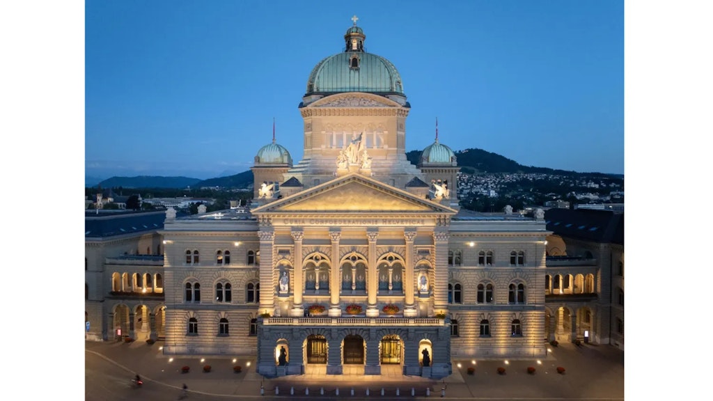 Vista esterna del Palazzo federale a Berna al crepuscolo, edificio neoclassico illuminato con cupola centrale e ali laterali, colline sullo sfondo.