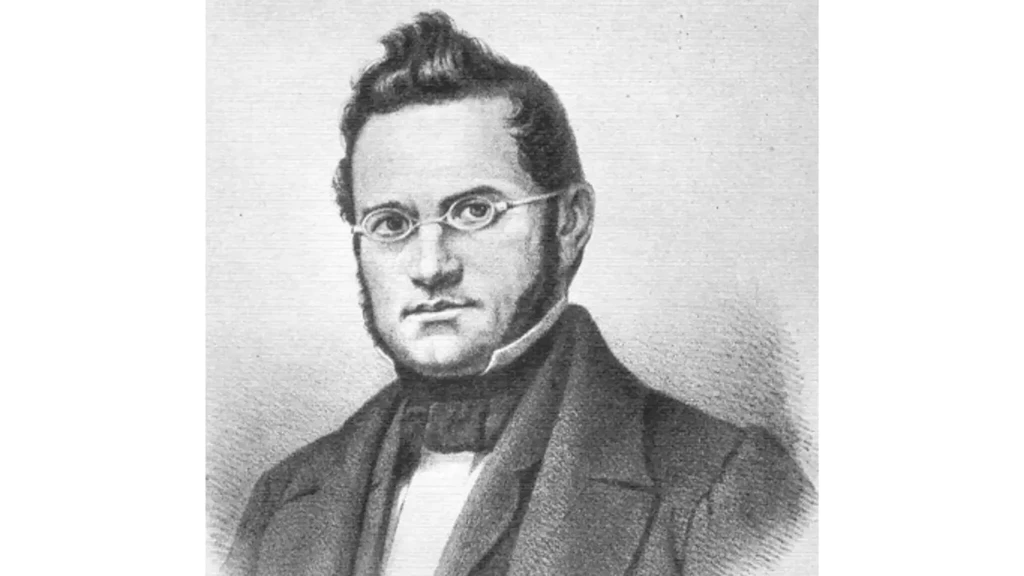 Schwarzweisses Porträt eines Mannes mit runder Brille, Backenbart und dunklem Anzug mit Fliege vor hellem Hintergrund.