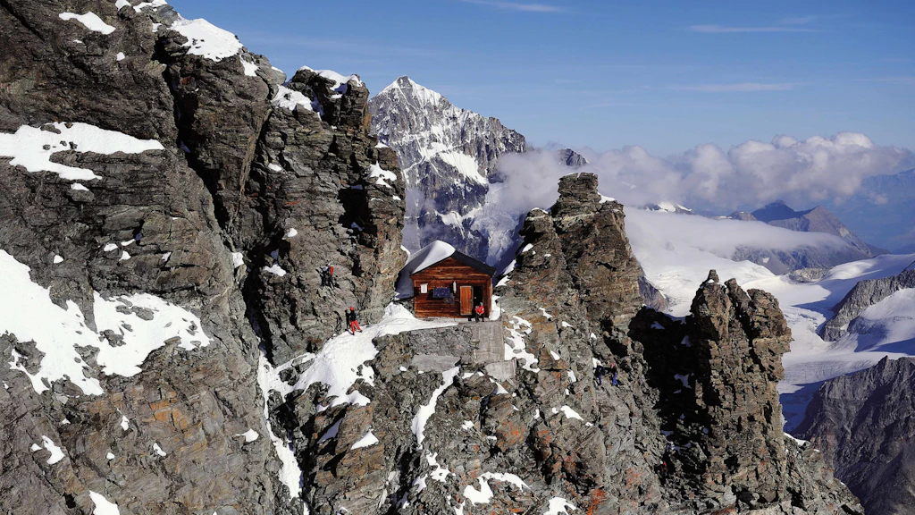 Kleine Berghütte aus Holz auf einem verschneiten Felsgrat in den Alpen, umgeben von Bergen und Gletschern.