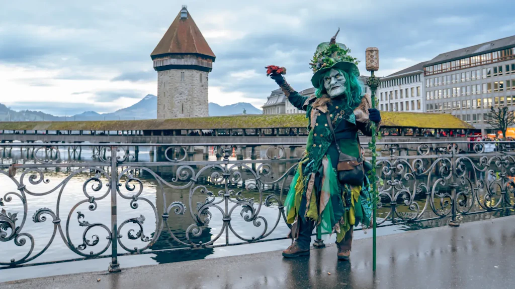 In Grün kostümierte Person mit Maske und geschmücktem Hut an einem Geländer am Wasser, mit Turm, gedeckter Brücke und Gebäuden im Hintergrund.