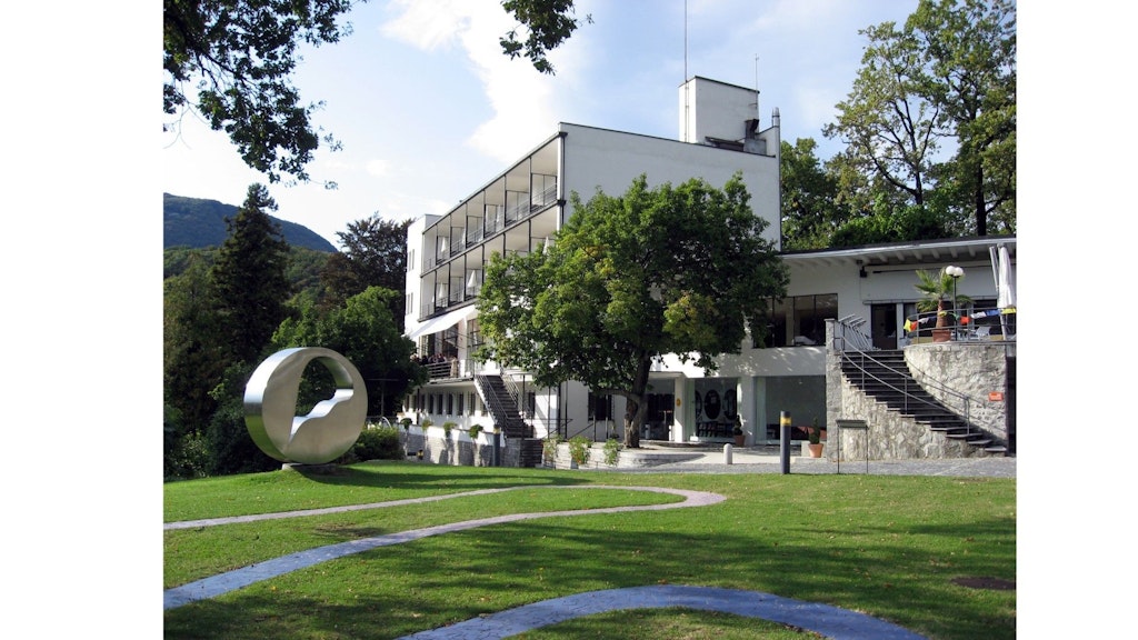 Modernes mehrstöckiges Gebäude mit Balkonen, umgeben von Bäumen, mit einer kreisförmigen Metallskulptur im Garten im Vordergrund. 