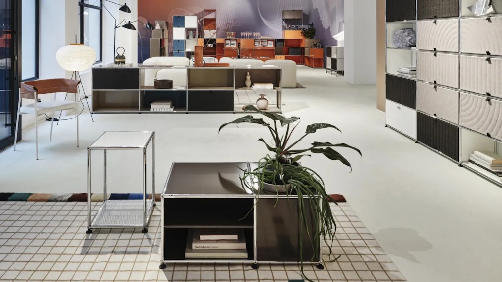 Innenraum eines Showrooms mit modularen USM-Möbeln, farbigen Regalen, Stuhl, Lampen und Pflanzen auf hellem Boden und kariertem Teppich.