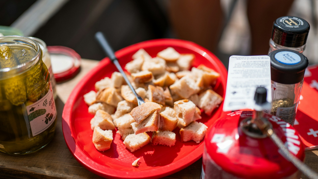 Roter Teller mit Brotstücken und Gabel, daneben ein Glas mit Essiggurken und Gewürzstreuer auf einem Holztisch.
