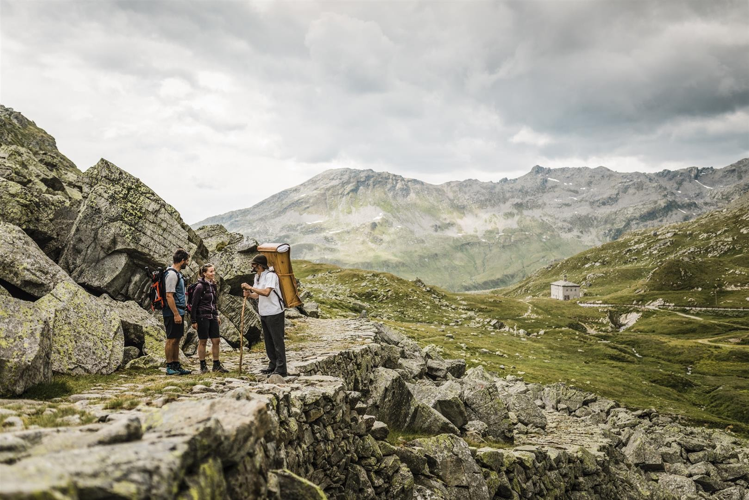 Drei Wanderer stehen auf einem felsigen Pfad in einer bergigen Gegend mit grünen Hügeln und wolkenverhangenen Bergen im Hintergrund.
