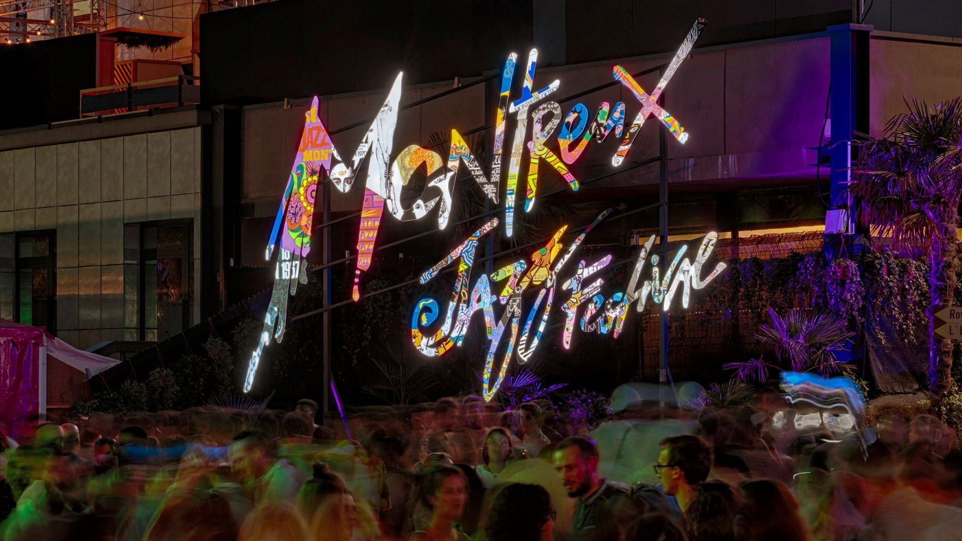Leuchtendes Schild „Montreux Jazz Festival“ mit bunten Buchstaben auf Metallgestell, verschwommene Menschenmenge im Vordergrund vor beleuchteten Industriegebäuden bei Nacht.
