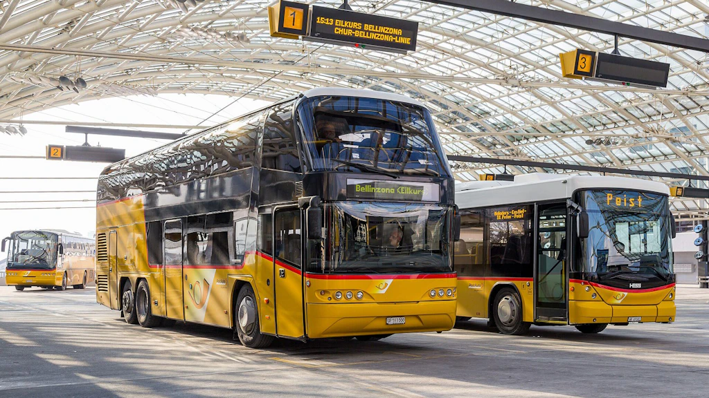 Überdachte Busstation mit mehreren gelb-roten Postautos in Reih und Glied unter einem Metallbau und hängenden Anzeigetafeln für die Bussteige.