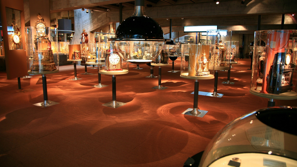 Innenansicht eines Ausstellungsraums mit dekorativen Uhren unter Glasglocken auf Ständern, rotem Teppichboden und sichtbarer Betonstruktur.