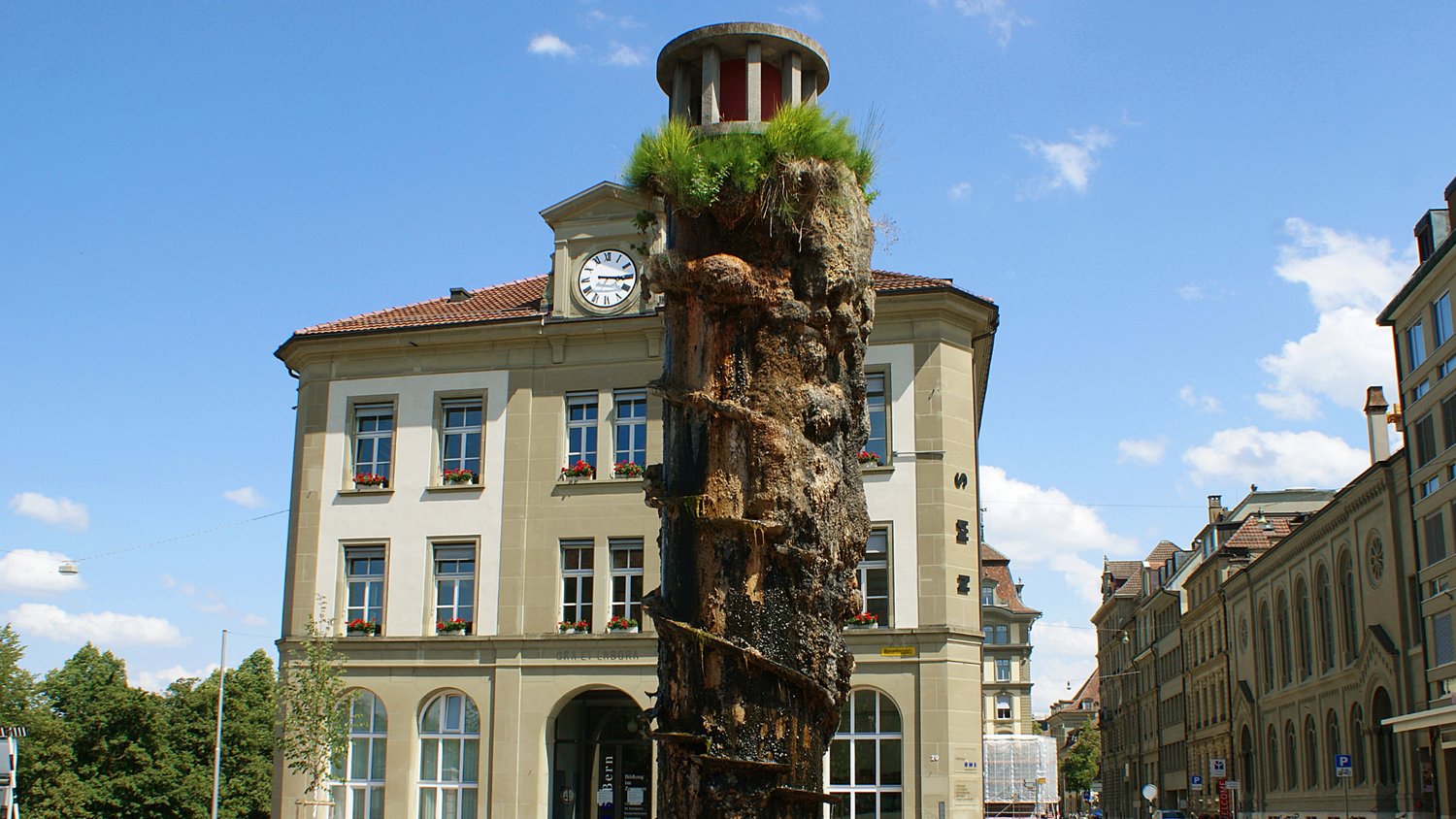 Brunnen mit einer organischen Säule und Pflanzen oben, vor einem historischen Gebäude in Bern, unter teilweise bewölktem blauem Himmel.