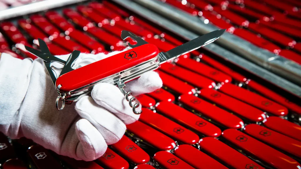 Mit Handschuh gehaltenes rotes Schweizer Taschenmesser mit ausgeklappten Werkzeugen über Reihen identischer roter Messer auf einer Metallfläche.