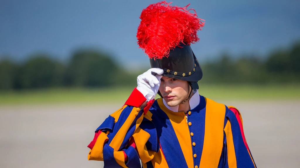 Ein Schweizer Gardist in blau-gelb-roter Uniform grüsst, indem er seinen schwarzen Helm mit rotem Federbusch mit seinem weissen Handschuh berührt, im Freien bei klarem Wetter.
