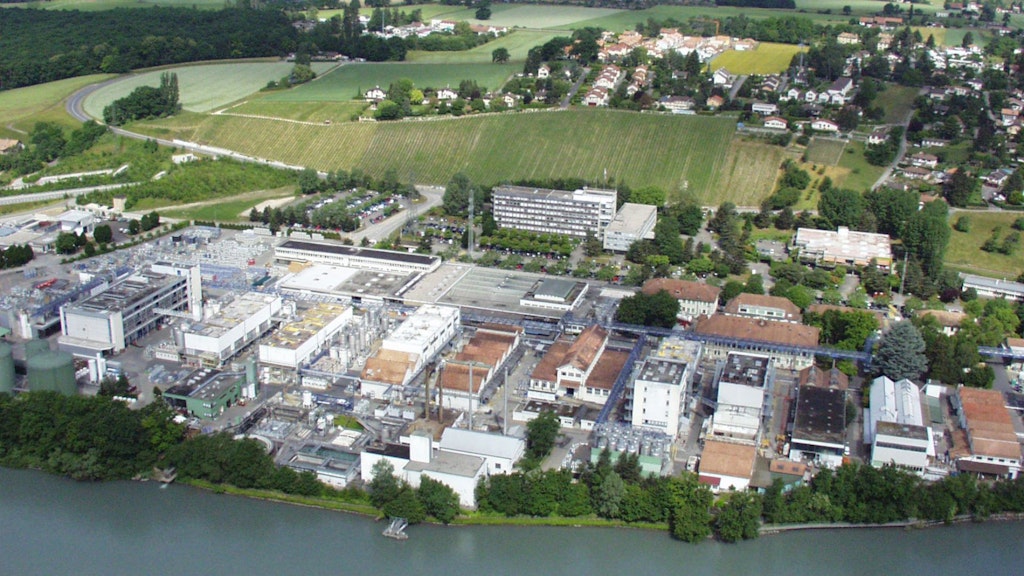 Luftaufnahme einer Industrieanlage an einem Flussufer mit mehreren Gebäuden, Parkplätzen, Weinreben und einem Wohngebiet im Hintergrund. 