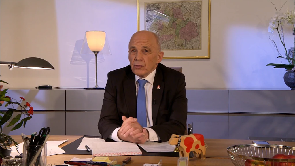 Mann im Anzug an einem Schreibtisch sitzend, mit gefalteten Haenden, Dokumenten, einer Lampe, gerahmter Karte an der Wand und einer Holzkuh auf dem Tisch.