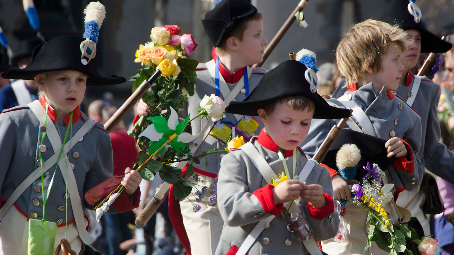 Kinder in historischen Uniformen mit pomponverzierten Hüten tragen Holzgewehre und Blumen, während sie an einer Freiluftveranstaltung teilnehmen.