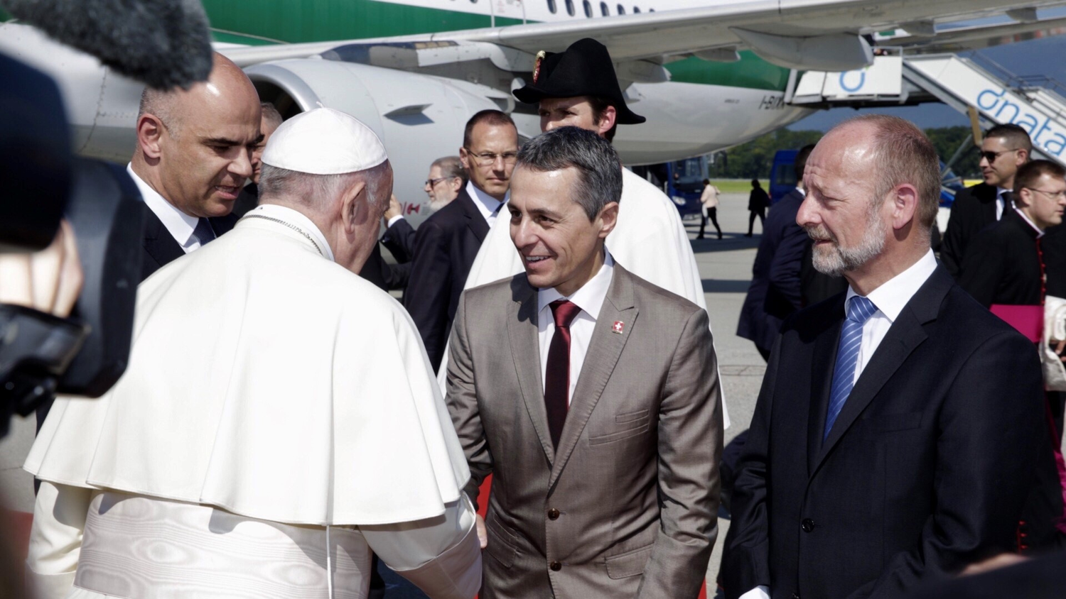 Papst Franziskus schüttelt einem Mann im hellen Anzug auf dem Rollfeld einesFlughafens die Hand, umgeben von mehreren Beamten in Anzügen, mit einem Flugzeug im Hintergrund.