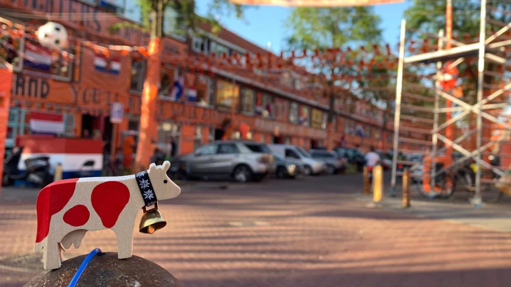 Hölzerne Kuhfigur mit roten Flecken und Glocke auf einem Metallpfosten, mit Strasse, geschmückten Gebäuden und unscharfen Autos im Hintergrund.