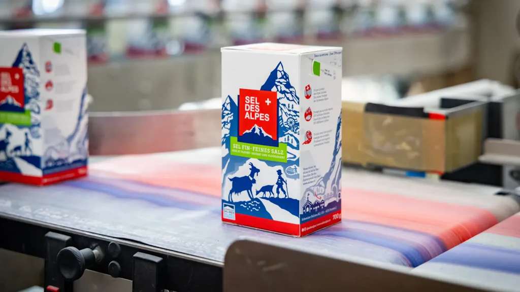 Packung Alpen-Salz auf einem Förderband in der Verpackung, mit Maschinen und weiteren Verpackungen im unscharfen Hintergrund.
