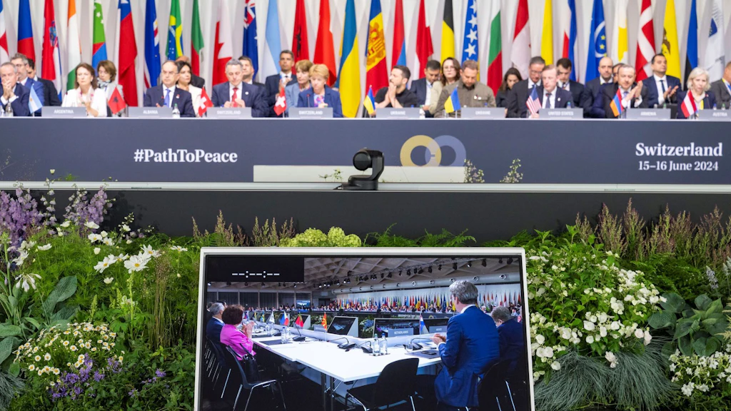 Das Bild zeigt die internationale Konferenz #PathToPeace in der Schweiz 2024, mit hochrangigen Delegierten vor Nationalflaggen und einem Monitor, der eine Videokonferenz zeigt.