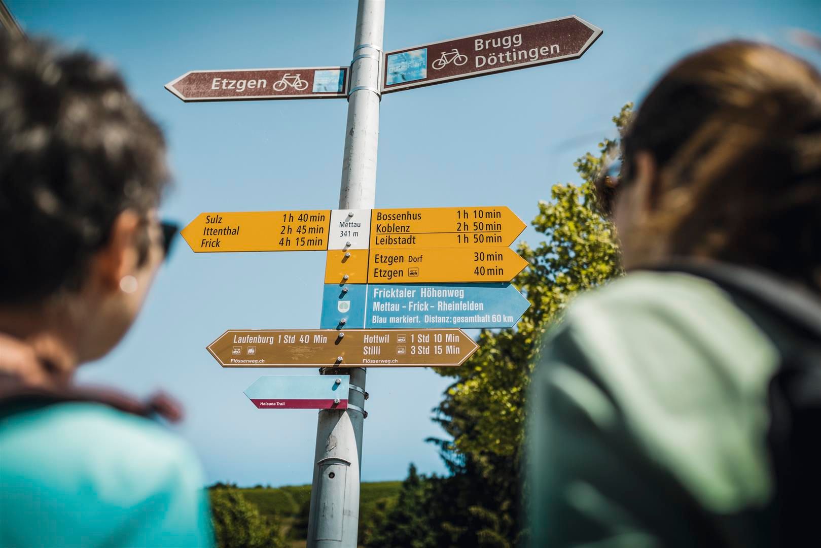 Wanderwegweiser mit bunten Pfeilen, Zielen und Gehzeiten an Metallmast, zwei unscharfe Wanderersilhouetten im Vordergrund.