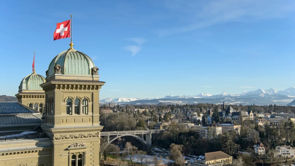 Das Bundeshaus in Bern mit einer wehenden Schweizer Flagge auf der Kuppel und schneebedeckten Alpen im Hintergrund unter blauem Himmel.