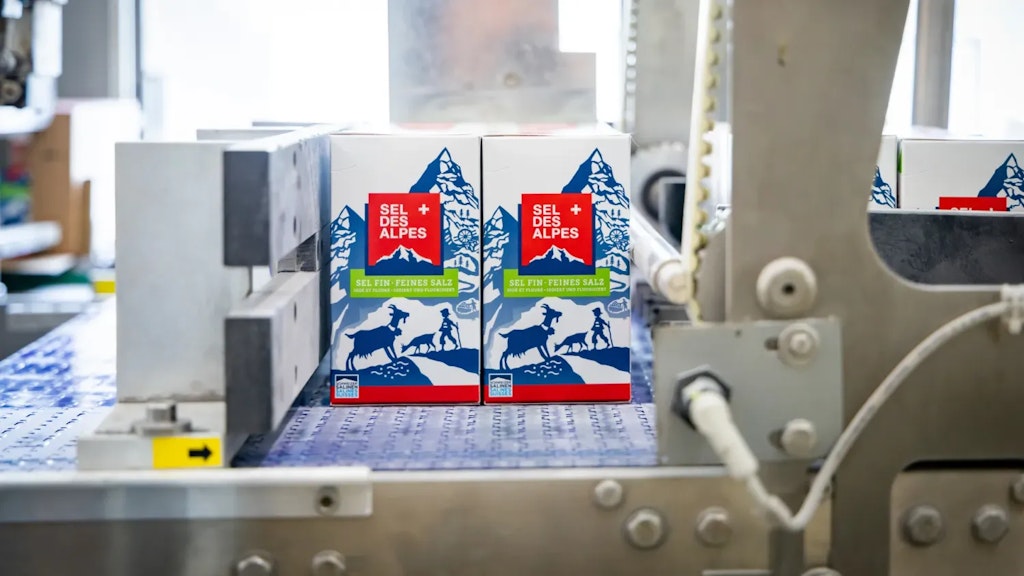Zwei Packungen Alpen-Salz nebeneinander auf einem Förderband, umgeben von Teilen industrieller Verpackungsmaschinen.