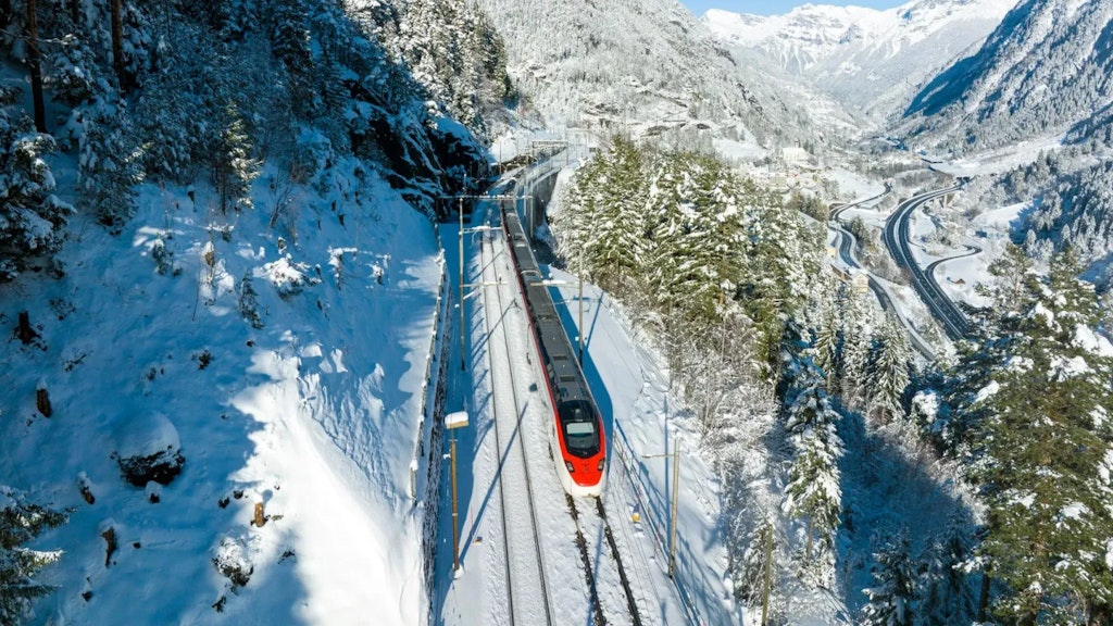 Rot-weisser Zug fährt auf einer verschneiten Bahnstrecke durch eine winterliche Berglandschaft, umgeben von Wäldern und Strassen im Tal.