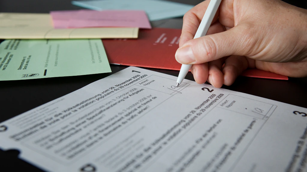 Hand hält Stift und schreibtauf einen gedruckten Stimmzettel, der auf einem Tisch liegt, im Hintergrund mehrere farbige Briefumschläge. 