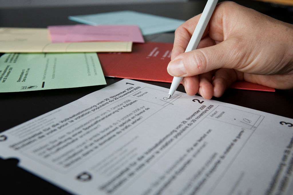 Hand hält Stift und schreibtauf einen gedruckten Stimmzettel, der auf einem Tisch liegt, im Hintergrund mehrere farbige Briefumschläge. 