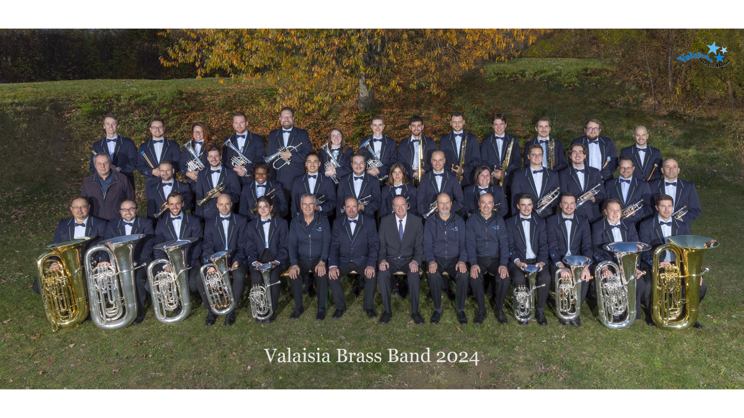 Gruppenfoto der Valaisia Brassband 2024: 40 Musiker, in dunklen Uniformen und auf drei Reihen in einem Park verteilt, halten Tuben, Posaunen, Hörner und Trompeten.