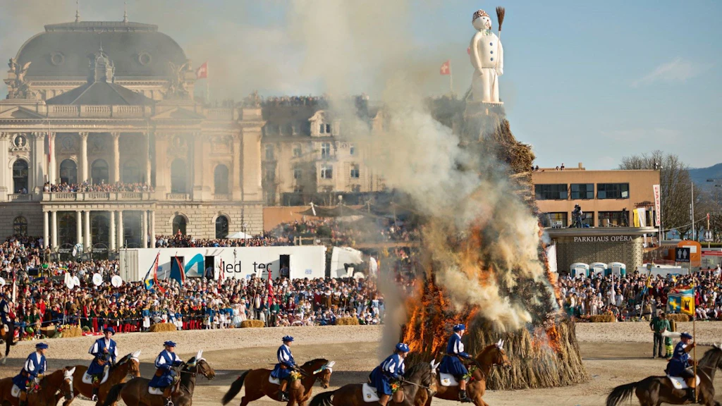 Reiter in traditioneller Kleidung galoppieren vor einem brennenden Holzhaufen mit einer weißen Figur an der Spitze, die Rauch abgibt, während einer Freiluftveranstaltung.