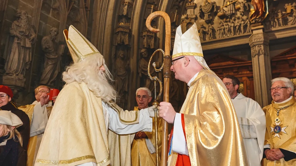  Zwei Männer in goldenen liturgischen Gewändern geben sich in einer Kirche die Hand, umgeben von Geistlichen.