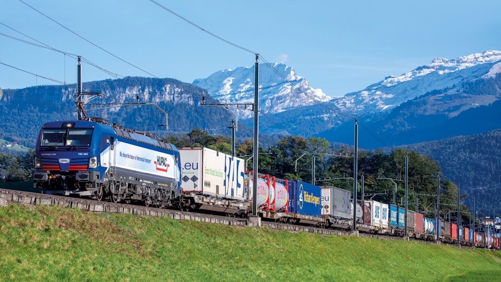 Ein Güterzug zieht mehrere Containerwagen mit verschiedenem Frachtgut durch eine Berglandschaft.