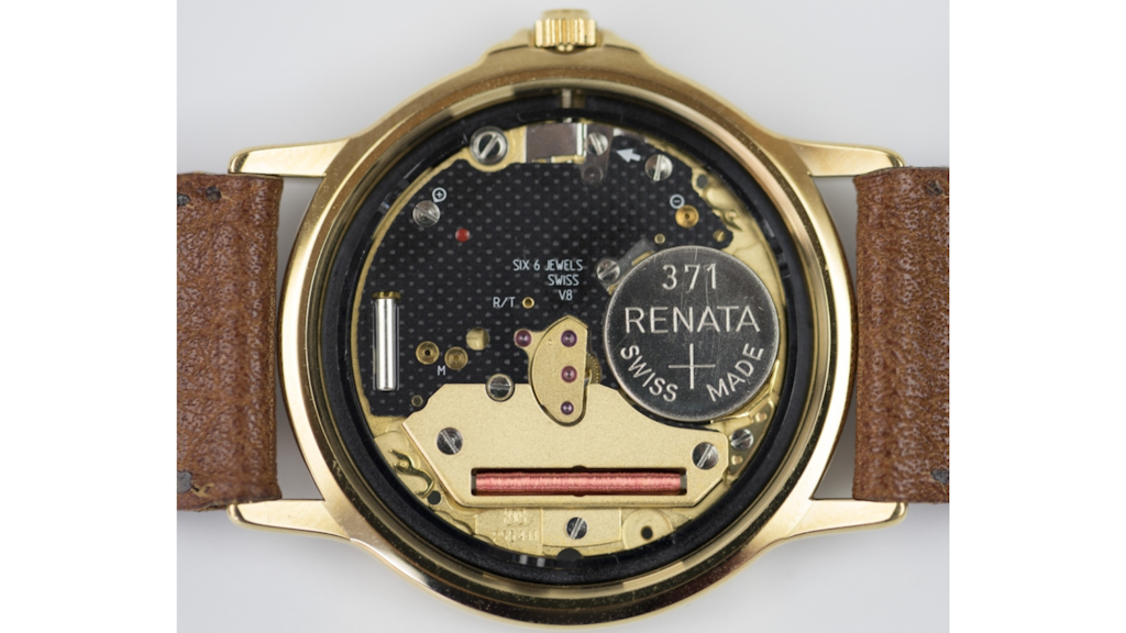Nahaufnahme des Inneren einer geöffneten Quarzuhr mit Renata 371-Batterie, Schaltkreisen, Spule und mechanischen Bauteilen in einem goldenen Gehäuse mit Lederband.