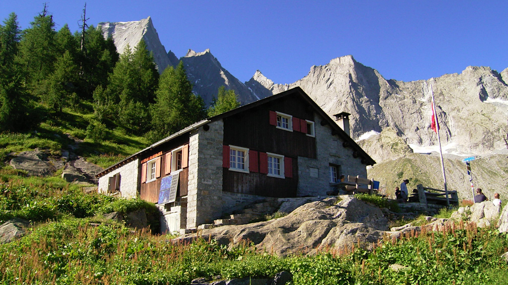 Berghütte aus Stein und Holz mit roten Fensterläden auf einer grünen Hanglage unter felsigen Gipfeln.