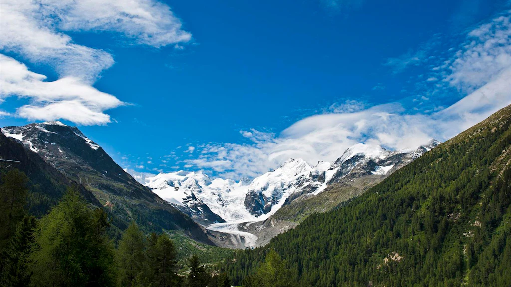 View of an Alpine valley with forests and snow-capped peaks against a blue sky with a few clouds.