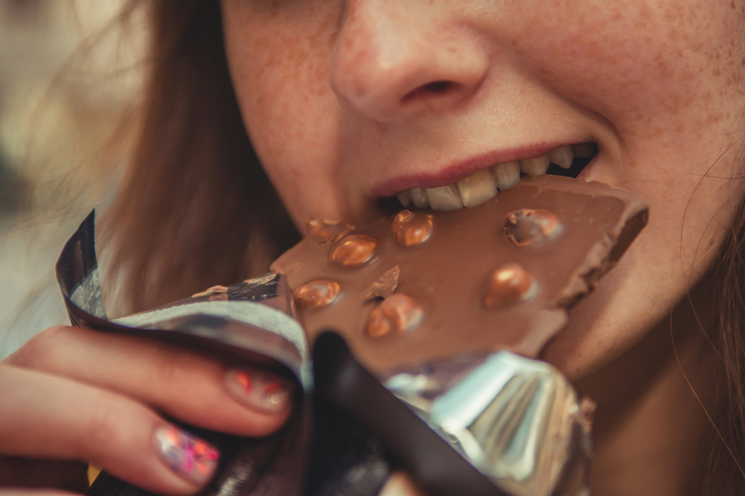 Close-up of a woman biting into a hazelnut chocolate bar, partly wrapped in silver foil.