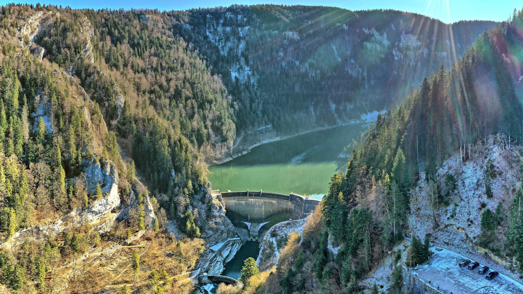 Aerial view of a curved dam in a narrow, wooded gorge, with a green reservoir behind it, steep rocky cliffs and a bright sun glinting on the right.