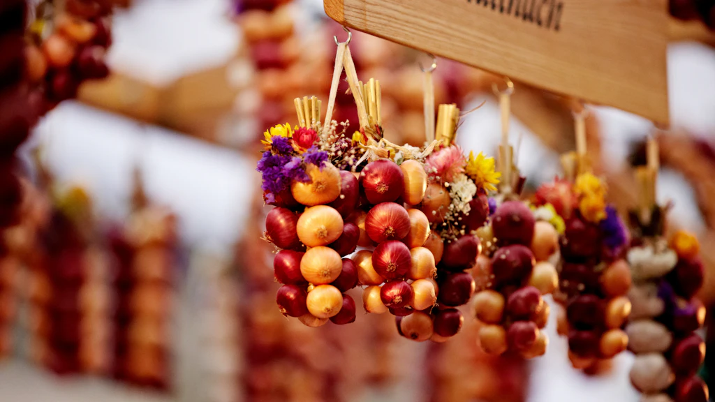 Garlands of red and yellow onions decorated with flowers, hanging at a market, with a wooden sign visible in the background.